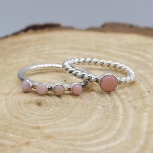 May include: Two silver rings with pink gemstones. One ring has a twisted band with a single, round pink stone. The other ring has a smooth band with four smaller pink stones. The rings are displayed on a wooden surface.