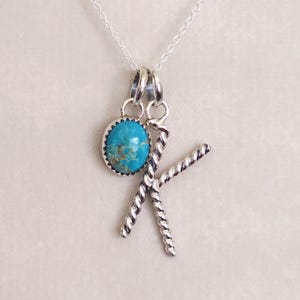 May include: A silver necklace featuring a pendant with a round, blue-green turquoise stone in a serrated bezel setting, paired with a separate silver charm shaped like the letter K with a twisted rope texture, both hanging from a silver chain.
