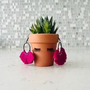Könnte beinhalten: Ein Terrakotta-Blumentopf mit gemalten Wimpern und rosafarbenen Quastenohrringen. Der Topf ist mit einer grünen Sukkulente bepflanzt.