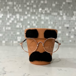 May include: A terracotta flower pot with a black mustache, eyebrows, and round glasses. The pot is sitting on a white countertop.