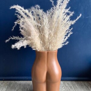 Op de afbeelding: Een bruine keramische vaas in de vorm van een menselijk lichaam, met een boeket gedroogd pampasgras erin. De vaas staat op een houten oppervlak.