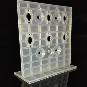May include: A clear, plastic display stand with a grid pattern and circular openings. The stand has a rectangular base and is designed to hold small items. The stand is made of a translucent material and is set against a black background.