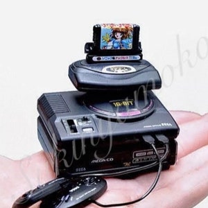 Può includere: Mini console Sega Mega Drive nera con una cartuccia di gioco con un personaggio anime femminile. La console ha un'etichetta a 16 bit e include due controller. L'immagine è su sfondo bianco.