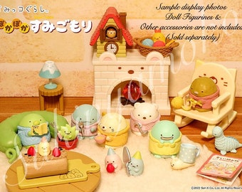 Op de afbeelding: Een verzameling Sumikko Gurashi-figuurtjes in verschillende poses, waaronder een pinguïn met een sjaal, een beer met een roze trui en een kat met een gele trui. De figuurtjes zijn gerangschikt rond een kleine taart en een open haard. De tekst "ぽ かすみごもり" is zichtbaar in de afbeelding.