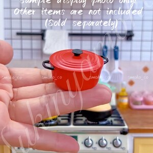 Puede incluir: Una olla de hierro fundido roja en miniatura con un asa y una tapa negras. La olla está siendo sostenida en la mano de una persona. El fondo muestra una cocina en miniatura con una estufa y otros artículos de cocina.