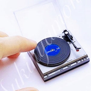 May include: A miniature silver record player with a black vinyl record on the turntable. The record has a blue label with the text "smoko_co" printed on it.