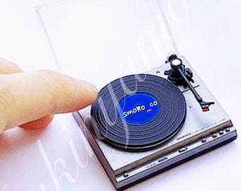 Può includere: Un giradischi argentato in miniatura con un disco in vinile nero sul piatto. Il disco ha un'etichetta blu con il testo "smoko_co" stampato su di esso.