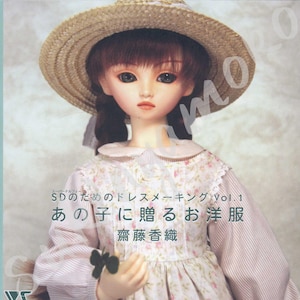 Può includere: Una bambola con un cappello di paglia e un vestito floreale. La bambola ha i capelli castani e tiene in mano un piccolo oggetto verde. Testo giapponese visibile sulla copertina di un libro, tra cui "SD no tame no doresume-kingu Vol.1".