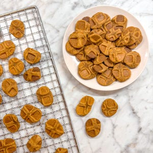 May include: A close-up of a wire rack with a batch of freshly baked pumpkin-shaped dog treats. The treats are a light brown color and have a slightly crispy texture. Some of the treats are on a white plate, while others are still on the rack.