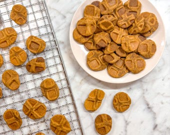 Peut inclure: Gros plan sur une grille en fil métallique avec un lot de friandises pour chiens en forme de citrouille fraîchement cuites. Les friandises sont de couleur brun clair et ont une texture légèrement croustillante. Certaines friandises sont sur une assiette blanche, tandis que d'autres sont encore sur la grille.