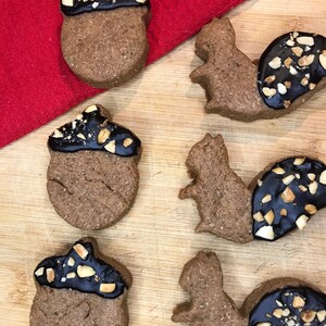 May include: Six decorated cookies on a wooden cutting board. Three cookies are shaped like acorns and three are shaped like squirrels. The cookies are decorated with chocolate icing and chopped nuts.