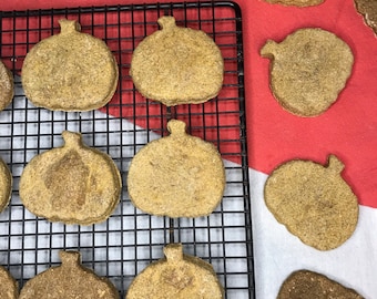 May include: A top-down view of pumpkin-shaped biscuits on a black wire rack and a red and white cloth. The biscuits are light brown with a stem detail. Some are on the rack, others are on the cloth.