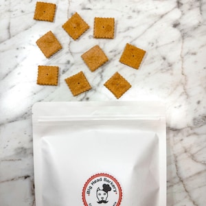 May include: A white bag with a red and white logo that says "Big Head Barkery Gourmet Dog Treats". There are 9 square-shaped dog treats scattered on a white marble surface.
