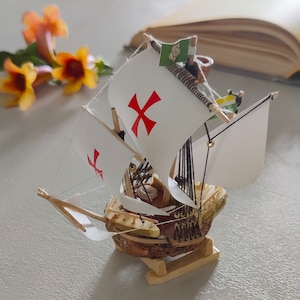 May include: A small wooden model ship with white sails and a red cross. The ship is on a wooden base and has a small green flag with a symbol on it.
