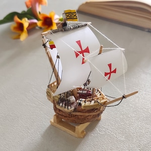 May include: A miniature wooden ship model with white sails and red crosses. The ship is decorated with a small flag with a yellow and black pattern. The ship is sitting on a white surface.