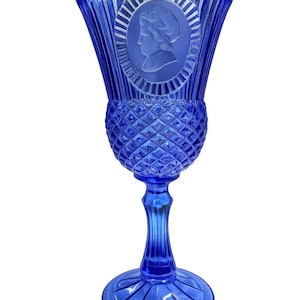 May include: A cobalt blue glass goblet with a detailed design. The upper portion features a cameo of a woman's profile within an oval frame. The goblet has a diamond-patterned midsection and a fluted stem, resting on a circular base.