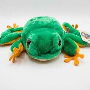 May include: A plush green frog toy with yellow accents on its feet and underside. The frog has large, black eyes and a tag attached to its side. The toy is designed to resemble a tree frog.