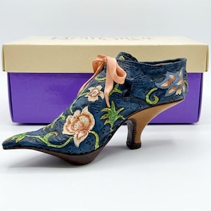 May include: A decorative shoe figurine in dark blue with a textured surface, featuring painted peach-colored floral patterns, green vines, and a small peach ribbon tied at the front. The shoe has a brown heel and sits in front of a box.