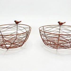 Può includere: Due cesti in filo metallico color rame con una struttura aperta simile a un nido. Ogni cesto a forma di ciotola presenta una piccola statuetta di uccello solida appollaiata sul bordo. La struttura in filo crea un motivo incrociato sul corpo.