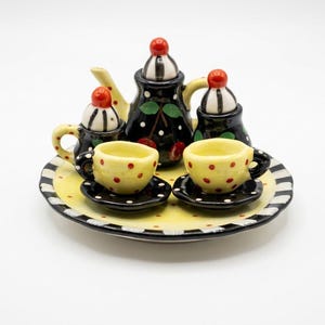 Può includere: Un set da tè in ceramica in miniatura composto da una teiera nera con motivi a ciliegie rosse, due tazze gialle a pois rossi su piattini neri, una zuccheriera, una lattiera e un grande vassoio giallo con bordo a strisce bianche e nere.