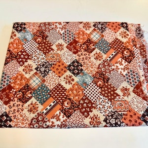 May include: A folded patchwork fabric featuring a variety of patterns and colors. The fabric is composed of numerous square patches in shades of red, orange, brown, and cream, with floral, plaid, and geometric designs. The edges of the fabric are frayed.