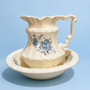 May include: A cream-colored ceramic pitcher and matching basin set featuring a scalloped rim and a decorative handle. The exterior of both pieces is adorned with a blue floral pattern consisting of roses and leaves on a smooth glazed surface.