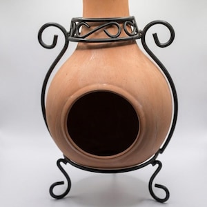 Könnte beinhalten: Ein Terrakotta-Kamin mit rundem Körper, einer großen kreisförmigen Öffnung, einem schmalen Hals und einem schwarzen Metallständer. Der Ständer ist mit dekorativen Schnörkeln versehen, die den Hals und den Boden des Tongefäßes umschließen.