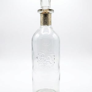 May include: A clear glass bottle with a decorative stopper. The bottle has a cylindrical shape with a slightly rounded base and a decorative embossed design. The stopper has a gold-colored band with an ornate pattern.