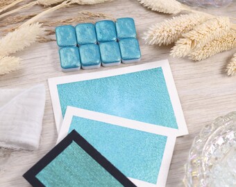 May include: A set of teal watercolor paint pans with a metallic sheen. The paint is displayed on a white card with a teal swatch. The card is on a light wood surface.