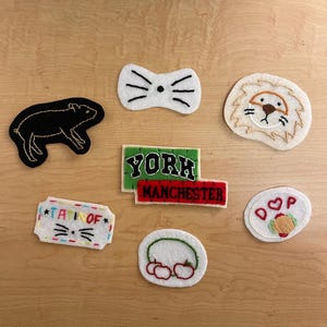 May include: A collection of embroidered patches in various shapes and designs. Includes a black rat, a white bow tie with whiskers, a lion's face, a patch with "YORK" and "MANCHESTER", a cat face with text, a pair of cherries, and a hot dog.