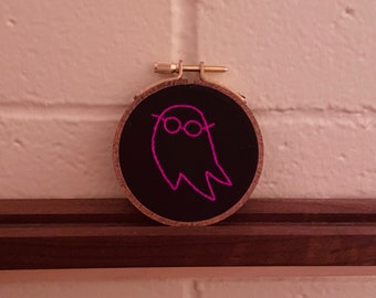 May include: A black embroidery hoop with a pink embroidered ghost wearing glasses. The ghost is outlined in pink thread.