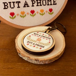 May include: A small wooden keychain with a white fabric circle featuring cross-stitch embroidery. The embroidery reads "Not a house but a home" with a small house design in the center. The keychain is on a wooden surface.