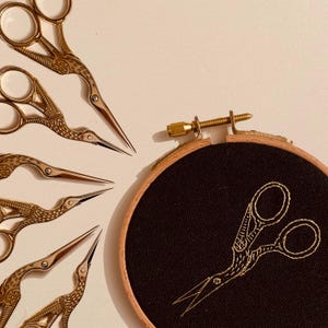 May include: A set of six gold-toned embroidery scissors with a bird design, arranged in a fan shape. A black embroidery hoop with a gold-toned embroidery design of a pair of scissors is in the background.