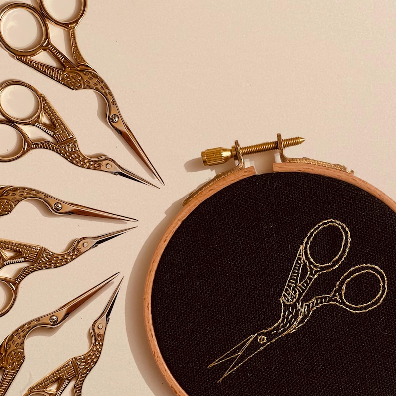 May include: A set of six gold-toned embroidery scissors with a bird design, arranged in a fan shape. A black embroidery hoop with a gold-toned embroidery design of a pair of scissors is in the background.