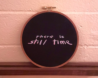 May include: A black embroidery hoop with the phrase "there is still time" embroidered in pink thread.