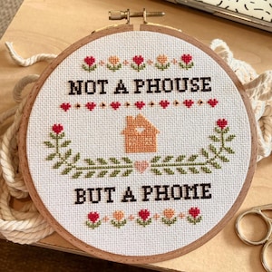 May include: A white embroidery hoop with a cross-stitch design that reads "Not a house but a home". The design features a small orange house with a heart in the window, surrounded by green leaves and red flowers.