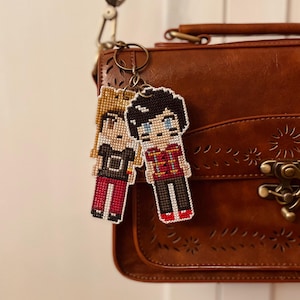 Op de afbeelding: Twee gepixelde sleutelhangers met figuren met zwart en blond haar, hangend aan een metalen ring. Eén figuur draagt een rood en zwart shirt, de ander een zwart shirt. De sleutelhangers zijn bevestigd aan een bruine leren tas.
