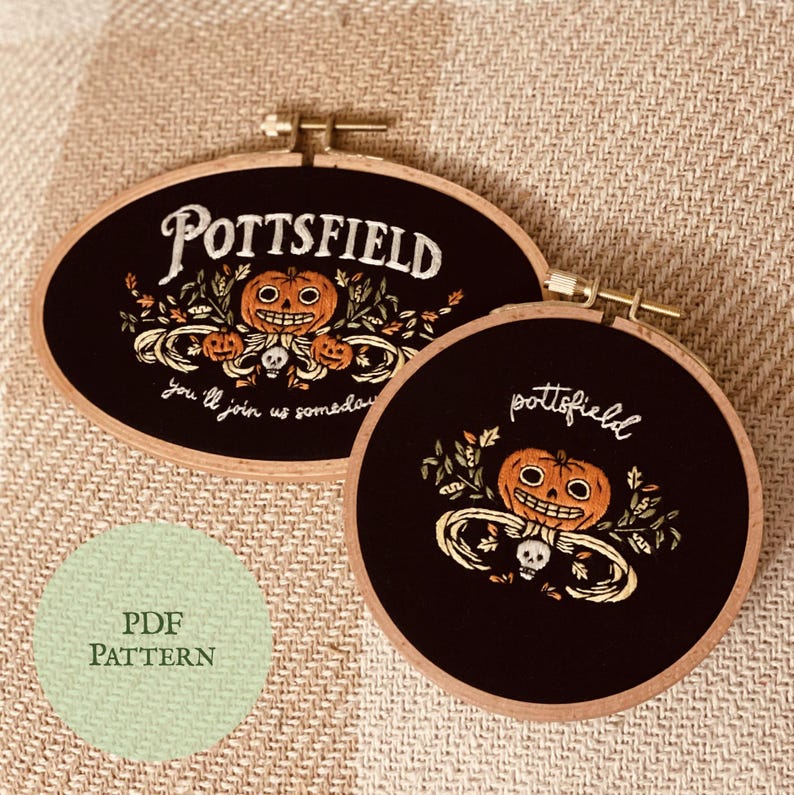 May include: Two black embroidery hoops with wooden frames. The hoops feature a Halloween-themed design with a jack-o'-lantern, skull, and leaves. The text "Pottsfield" and "You'll join us someday" are embroidered on the hoops.