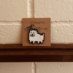 May include: A white and black pixelated dog embroidered on a brown card with the text "olivien gren" printed on it.