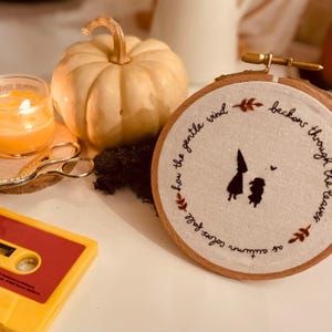 May include: Embroidered hoop art with a quote about autumn and a gentle wind. The quote reads "As autumn softly falls, how the gentle wind beckons through the trees". The hoop is brown wood with white fabric inside. There are two small figures embroidered in black on the fabric.