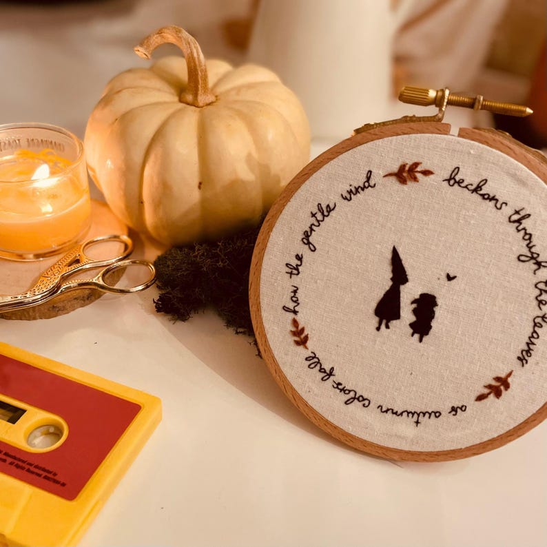 May include: Embroidered hoop art with a quote about autumn and a gentle wind. The quote reads "As autumn softly falls, how the gentle wind beckons through the trees". The hoop is brown wood with white fabric inside. There are two small figures embroidered in black on the fabric.