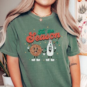 May include: Green t-shirt with the phrase "Tis the Season" in red and green lettering. The shirt features a cartoon cookie and milk carton holding hands and strung with Christmas lights. The shirt is a comfortable fit.