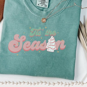 May include: A teal green t-shirt with a pink and white retro-style graphic that says "Tis' the Season" with a stylized Christmas tree.