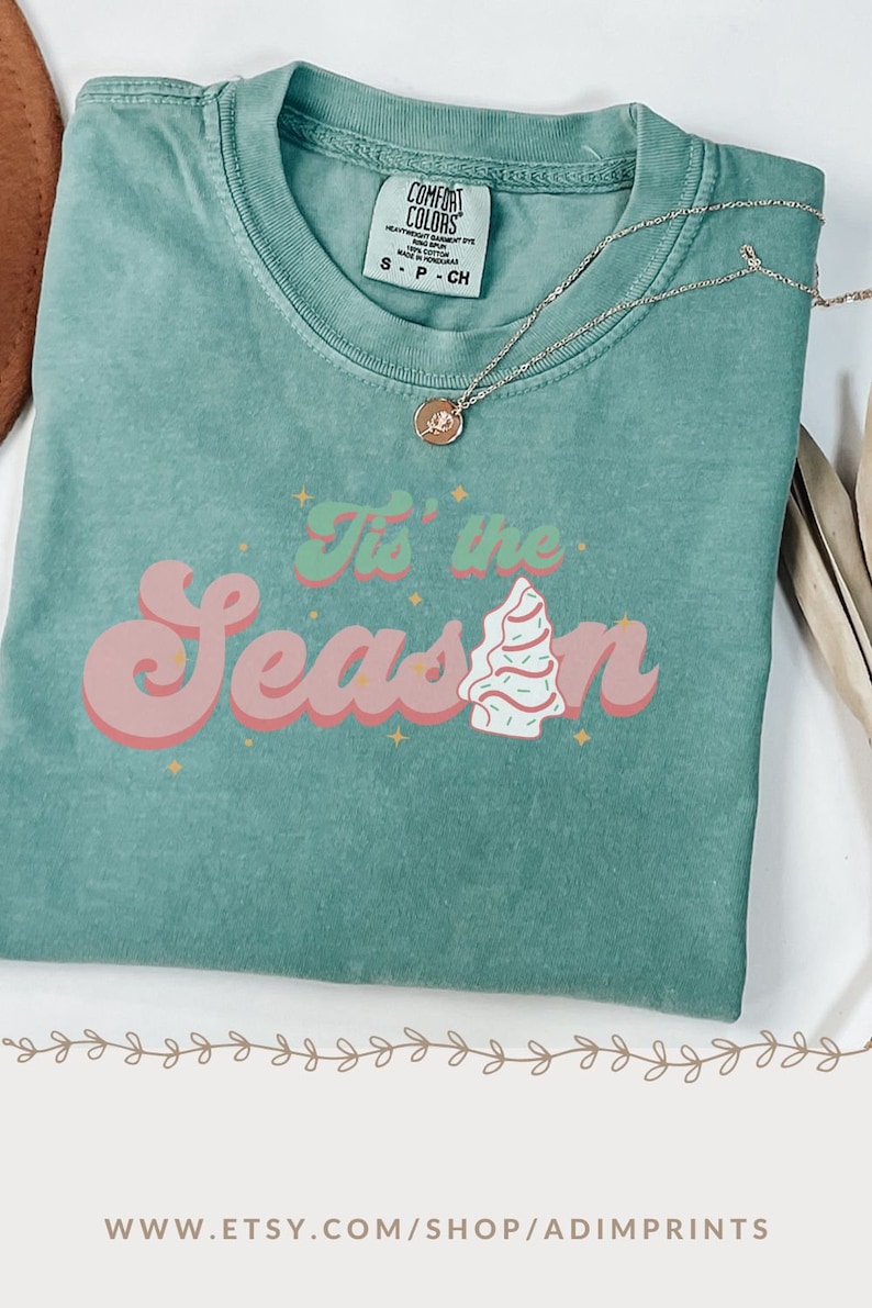 May include: A teal green t-shirt with a pink and white retro-style graphic that says "Tis' the Season" with a stylized Christmas tree.