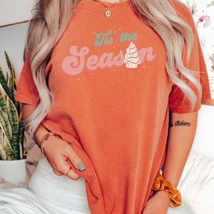 May include: A woman wearing a burnt orange t-shirt with the text "It's the Season" in a retro font with a stylized Christmas tree graphic.