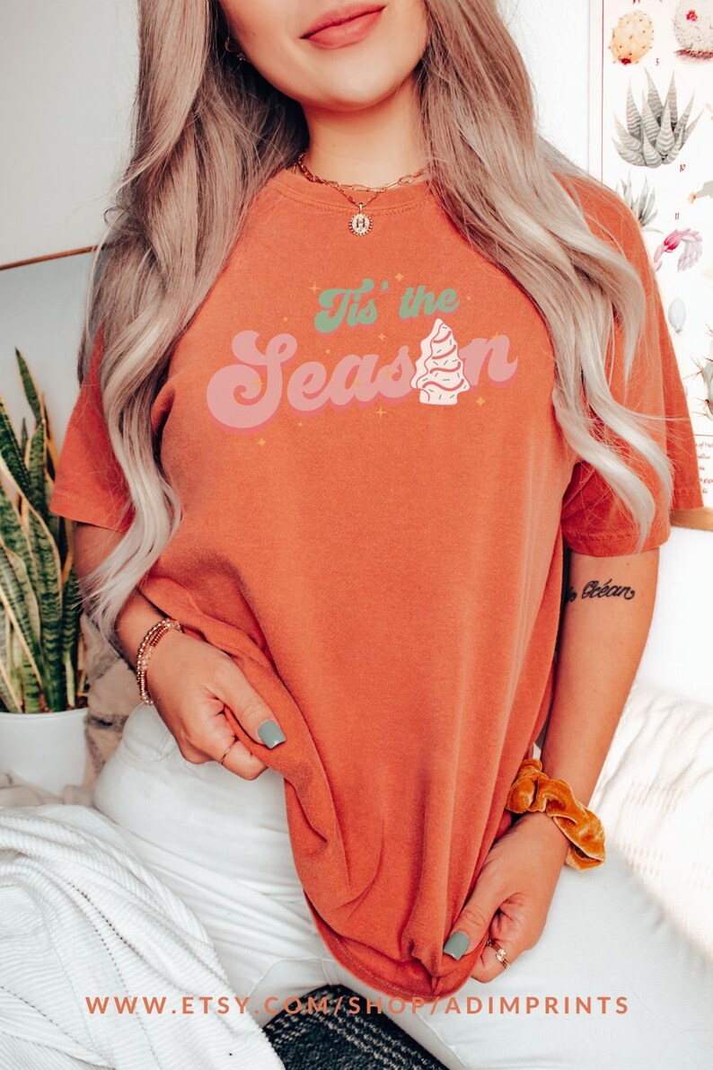 May include: A woman wearing a burnt orange t-shirt with the text "It's the Season" in a retro font with a stylized Christmas tree graphic.