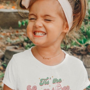 May include: A young girl wearing a white t-shirt with a pink and green "Tis' the Season" graphic. The girl is smiling and wearing a pink headband. The text "www.etsy.com/shop/adimprints" is at the bottom of the shirt.