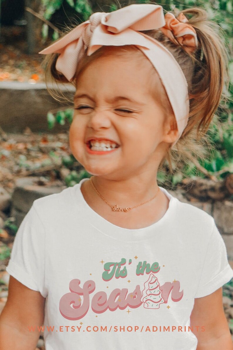 May include: A young girl wearing a white t-shirt with a pink and green "Tis' the Season" graphic. The girl is smiling and wearing a pink headband. The text "www.etsy.com/shop/adimprints" is at the bottom of the shirt.