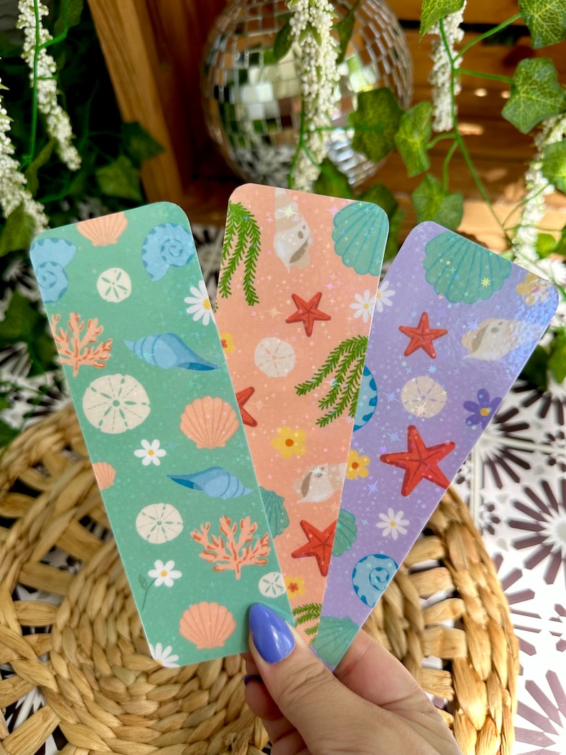 May include: Three colorful bookmarks with sea-themed designs. Each bookmark features illustrations of seashells, starfish, and coral in shades of green, peach, and purple. The bookmarks are held in a hand.