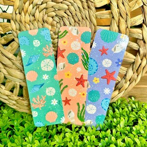 May include: Three colorful bookmarks with a sea life theme. Each bookmark features a different background color: mint green, peach, and lavender. The designs include seashells, starfish, and coral. The bookmarks are displayed on a woven background.
